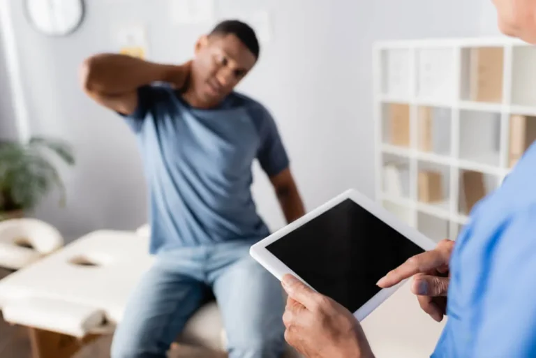 Man holding the back of his sore neck while a provider takes notes on a tablet during a consultation